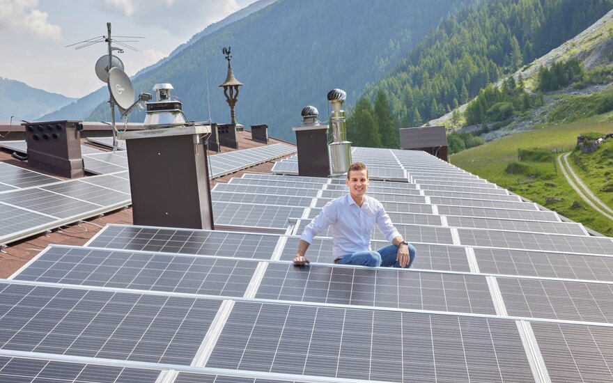 Ein junger Mann sitzt lächelnd auf einem Dach voller Solarpanels, umgeben von einer malerischen Berglandschaft.