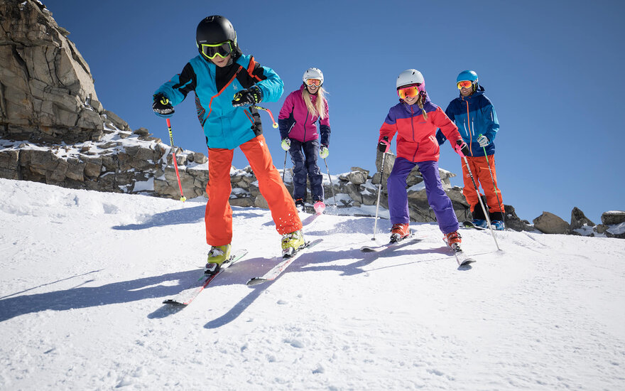 Eine Gruppe von vier Skifahrern fährt fröhlich einen schneebedeckten Hang hinunter, umgeben von felsigen Bergen und klarem Himmel.