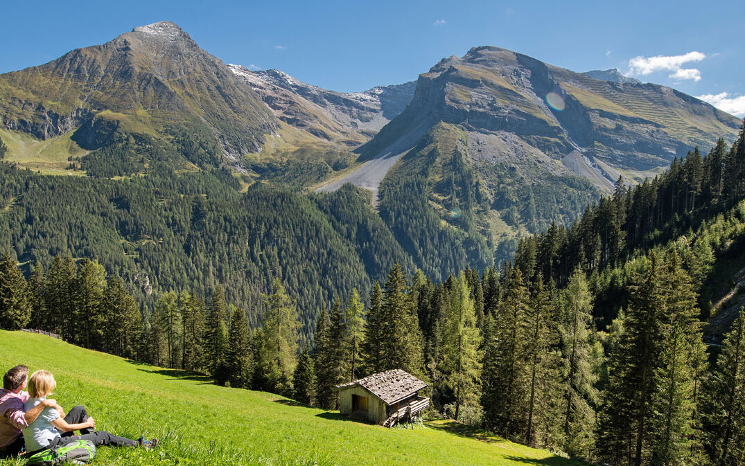 Ein Paar sitzt auf einer Wiese und genießt die atemberaubende Aussicht auf die Berge und den Wald im Hintergrund.