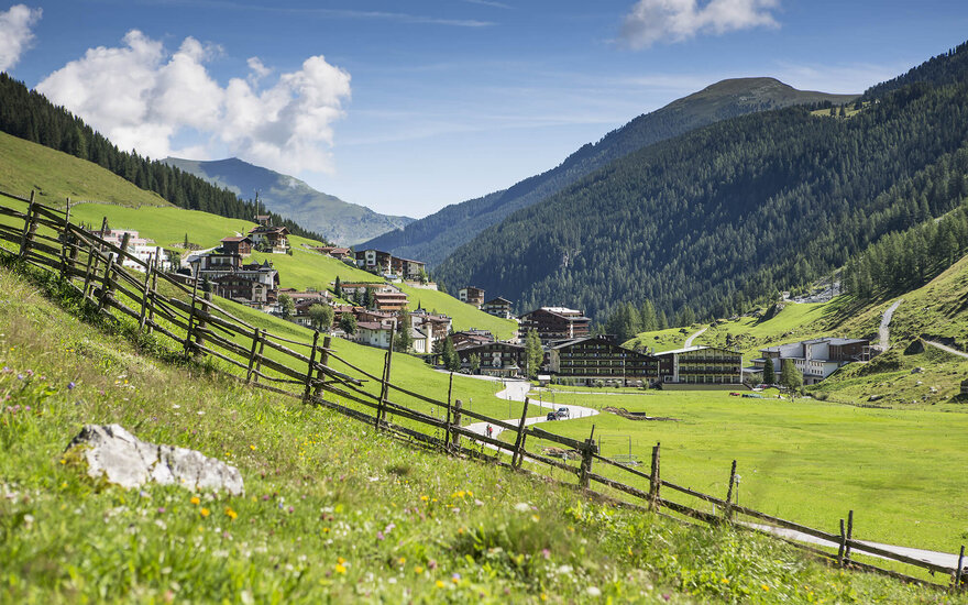 Eine malerische Berglandschaft mit grünen Wiesen und einem charmanten Dorf im Hintergrund, umgeben von hohen Bergen.