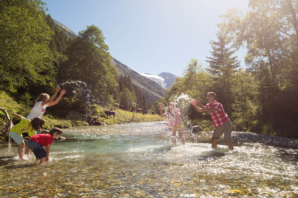 Eine fröhliche Familie spielt im klaren Wasser eines Flusses, während die Sonne strahlt und die Berge im Hintergrund sichtbar sind.