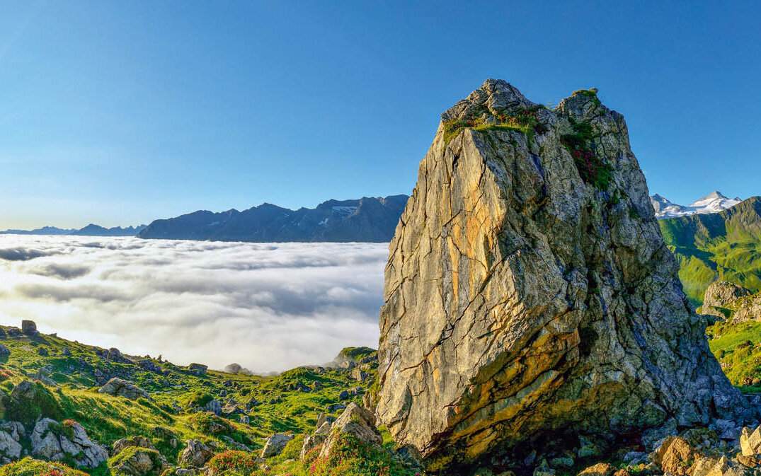 Die Sonne strahlt über die Berge, während große Felsen die Aussicht auf die darunter liegende Wolkenlandschaft rahmen.