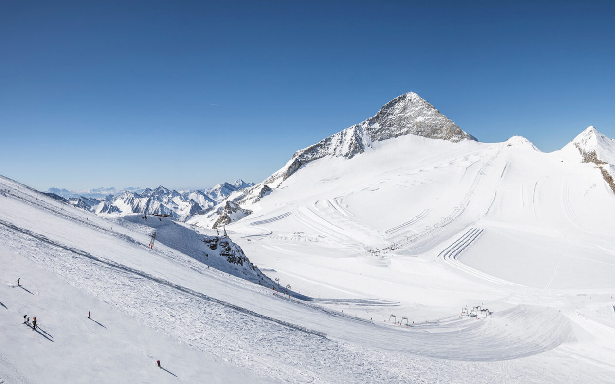 Eine weite, schneebedeckte Berglandschaft mit Skifahrern, die die Pisten hinunterfahren, unter einem strahlend blauen Himmel.