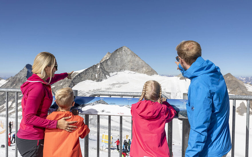 Eine Familie steht an einem Geländer und betrachtet die schneebedeckten Berge, während die Mutter auf einen Gipfel zeigt.