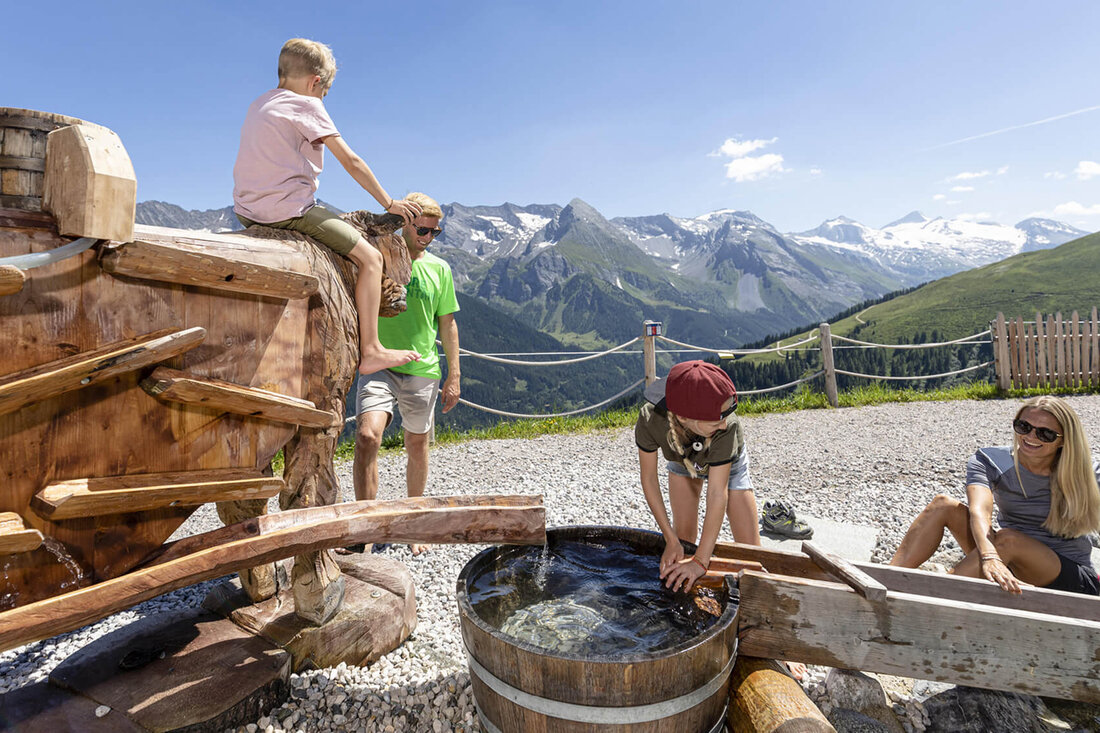 Eine Familie genießt einen sonnigen Tag in den Bergen, während Kinder an einem Wasserspielzeug spielen und lachen.