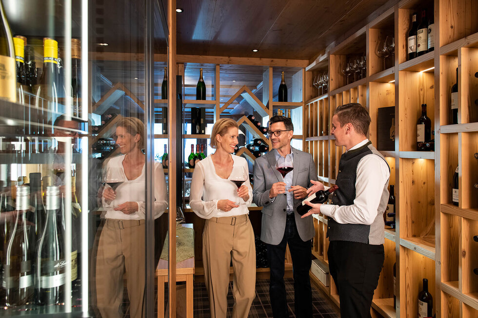 In einem eleganten Weinkeller unterhalten sich ein Paar und ein Sommelier über Weinauswahl, während sie Gläser in der Hand halten.