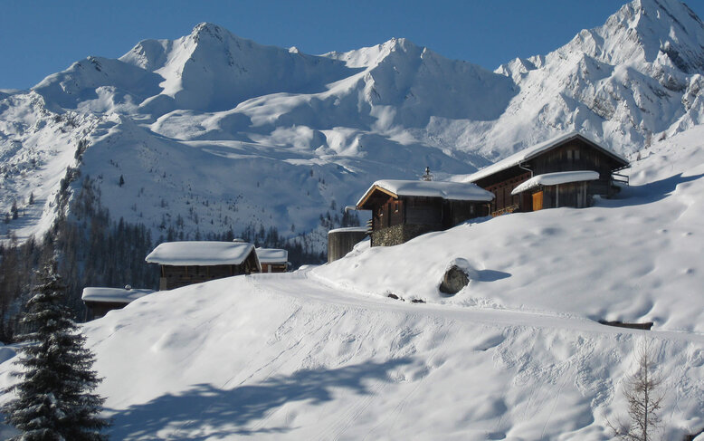 Schneebedeckte Hütten liegen malerisch vor einer beeindruckenden Berglandschaft unter klarem blauen Himmel.
