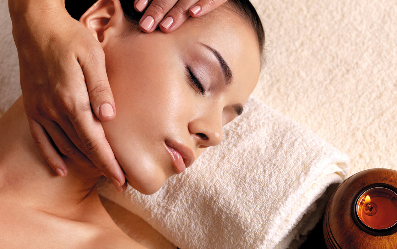 Eine entspannte Frau erhält eine Gesichtsmassage, während ihre Augen sanft geschlossen sind und eine Kerze im Hintergrund brennt.