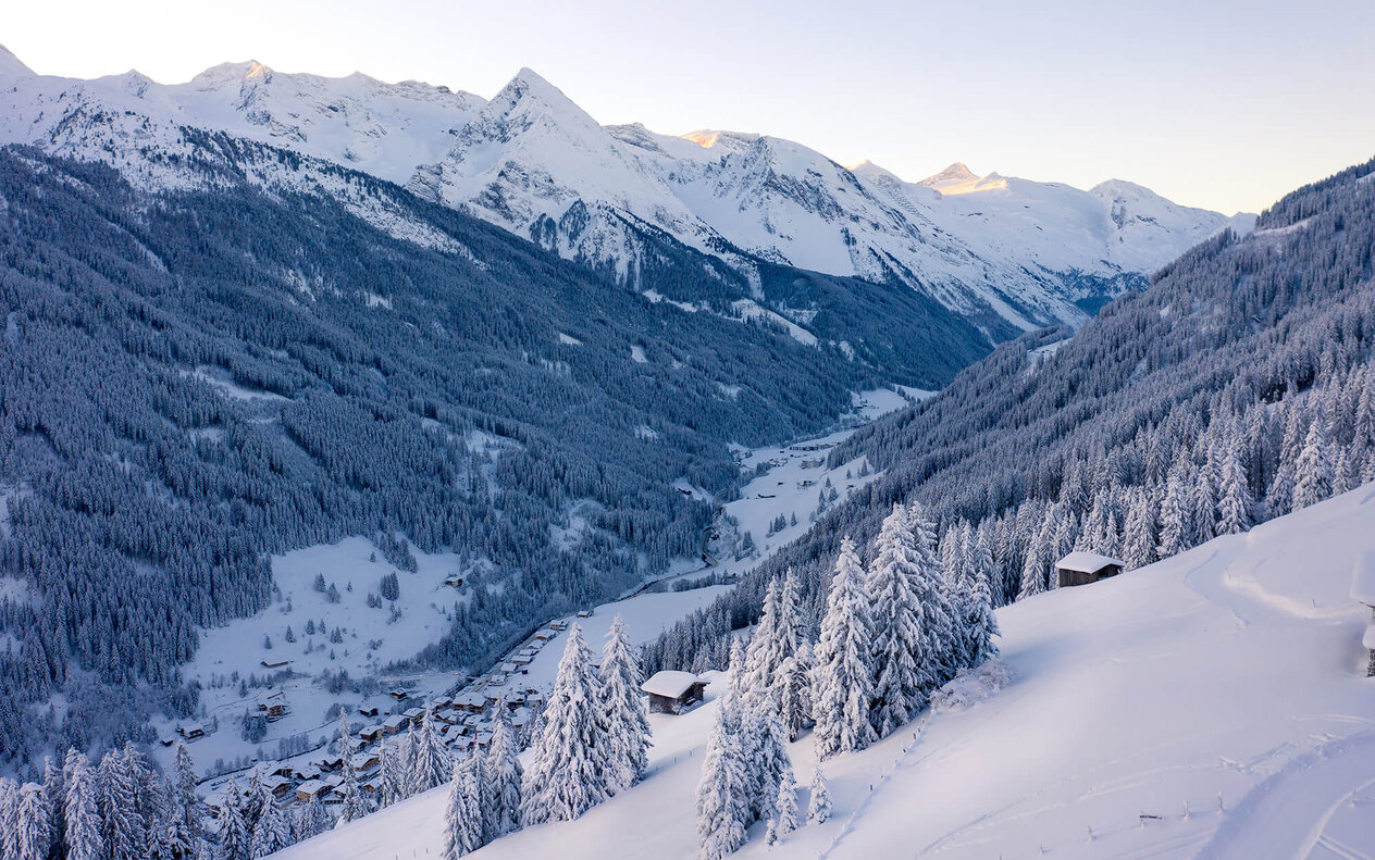 Eine verschneite Berglandschaft mit schneebedeckten Bäumen und einem kleinen Dorf im Tal, umgeben von majestätischen Gipfeln.