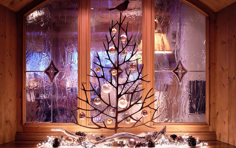 Ein stilvoller Weihnachtsbaum aus Ästen steht vor einem Fenster mit buntem, glitzerndem Licht und dekorativen Kugeln.
