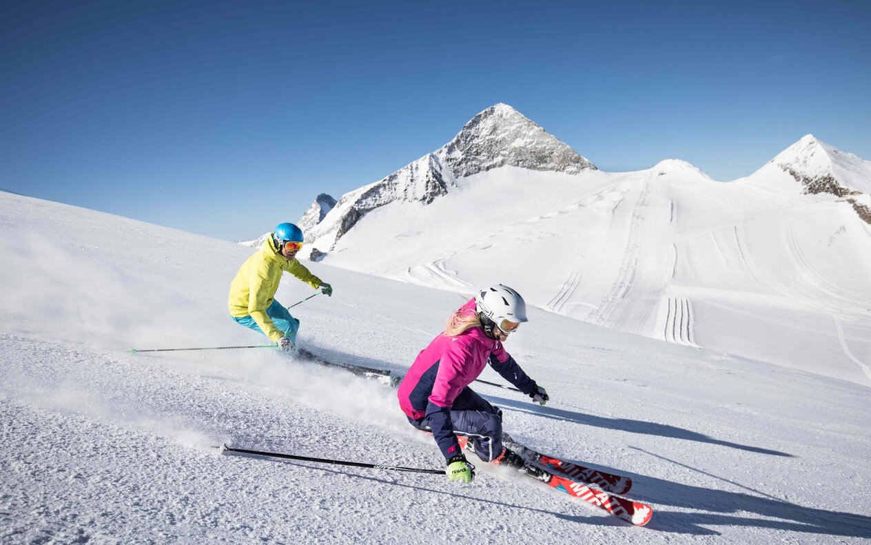 Zwei Skifahrer, ein Mann in gelber Jacke und eine Frau in pinker Jacke, rasen über die schneebedeckte Piste unter klarem Himmel.