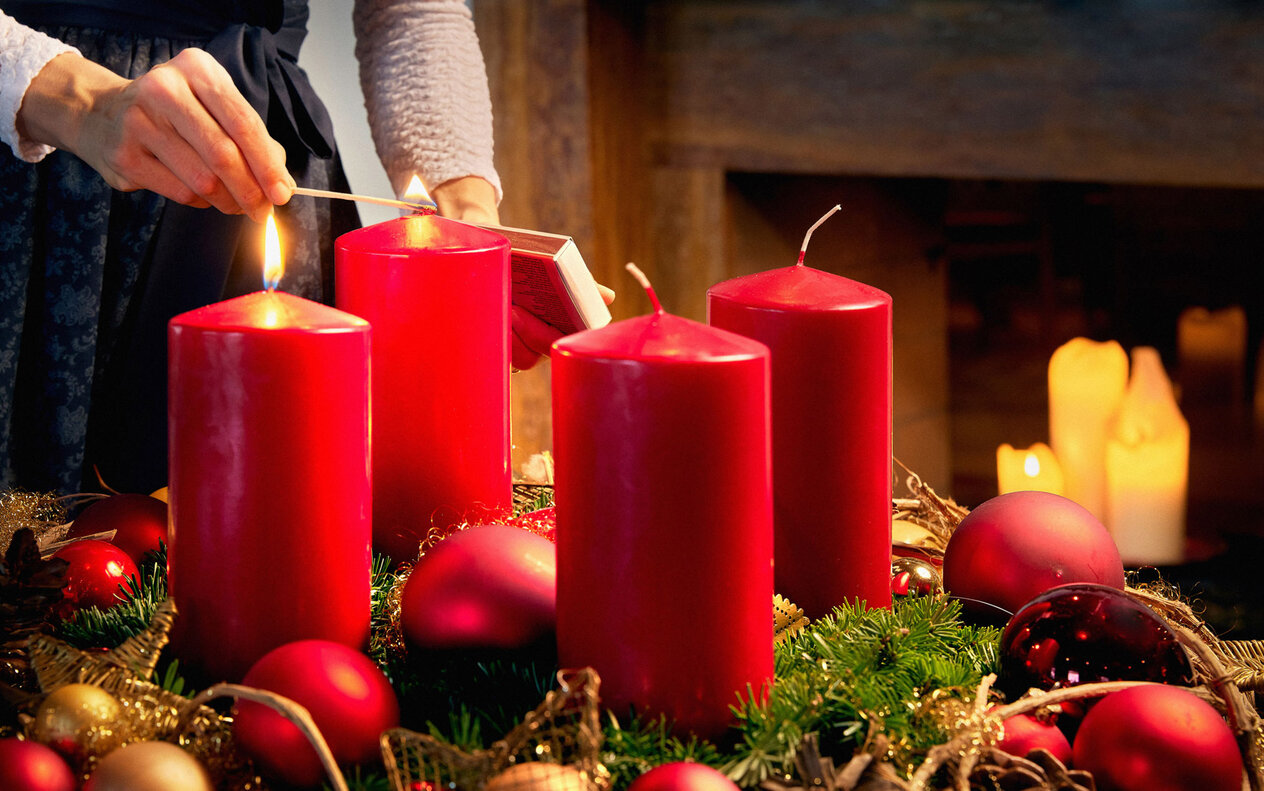 Eine Hand zündet eine rote Kerze an einem festlich dekorierten Tisch mit Weihnachtskugeln und Tannenzweigen an.