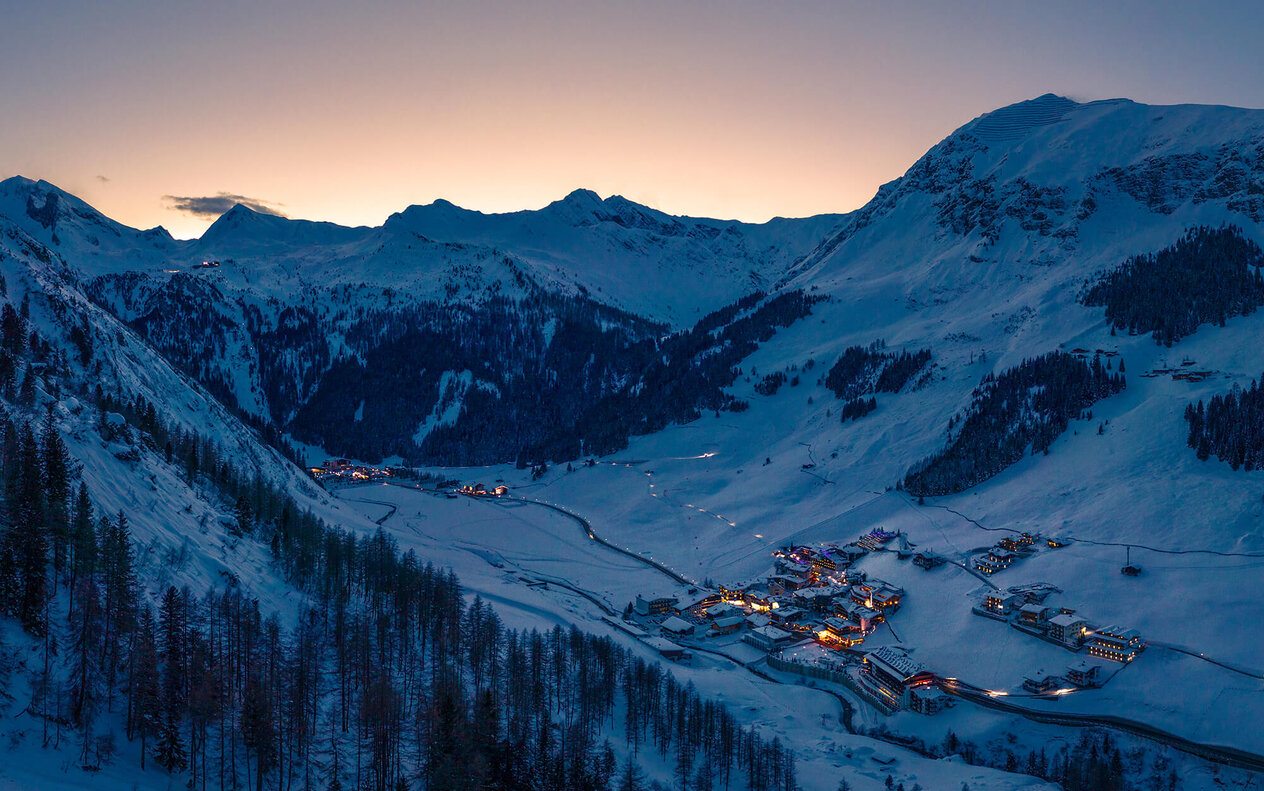 Die schneebedeckten Berge umgeben ein kleines, beleuchtetes Dorf in der Dämmerung, das eine friedliche Winteratmosphäre ausstrahlt.